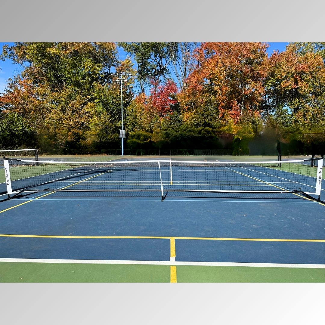 A blue and green outdoor pickleball court features the Playly Store Logo Portable Pickleball Net at its center, surrounded by autumn foliage in green, yellow, and red under a clear sky with sunlight casting shadows across the empty court.