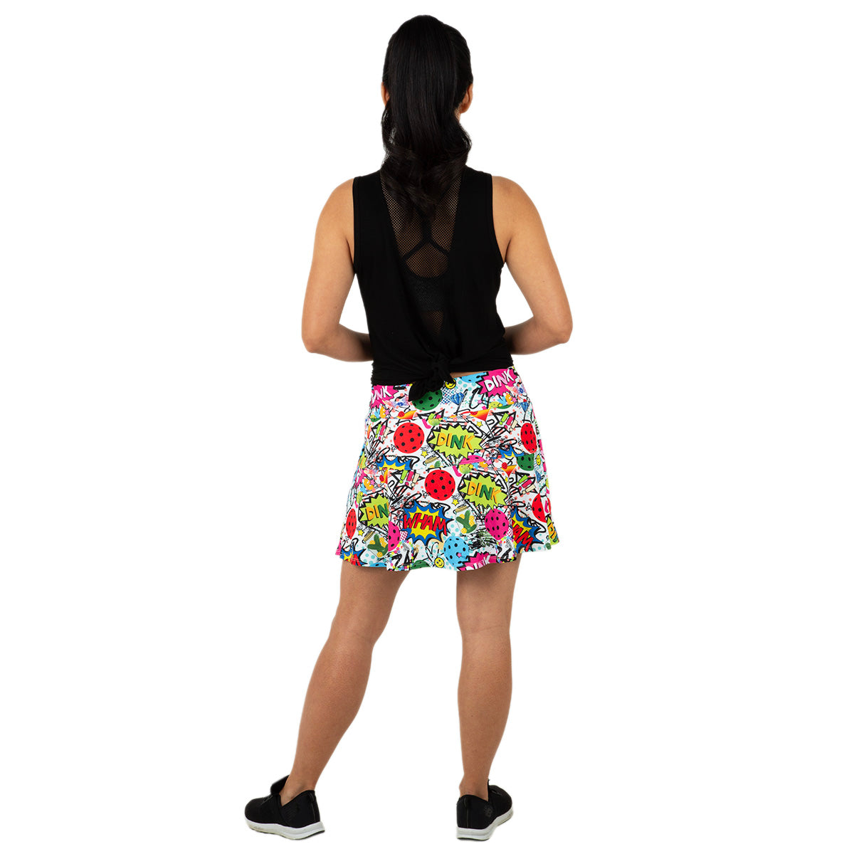 A person stands facing away, wearing the Queen of the Court Dink Dink Wham Swing Skirt with comic book-style prints and a sleeveless black mesh-back top. Their dark hair is in a ponytail, with black sneakers, against a white background.