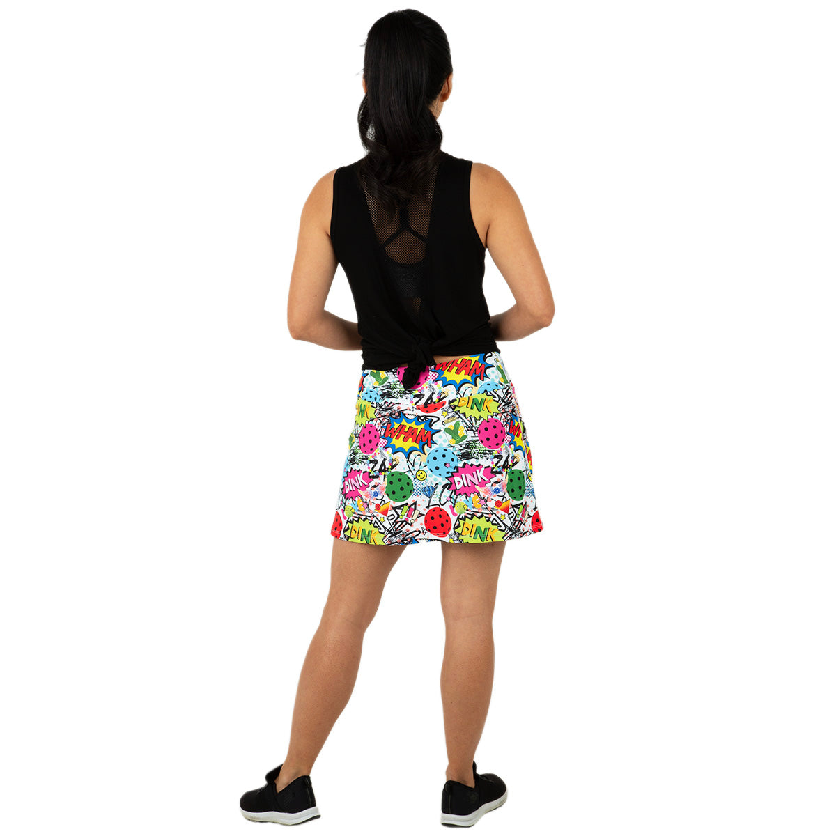 A woman with a ponytail faces away, wearing Queen of the Court’s Dink Dink Wham Endurance 15" Skirt featuring comic-style prints and a sleeveless black top, paired with black sneakers against a plain white background.