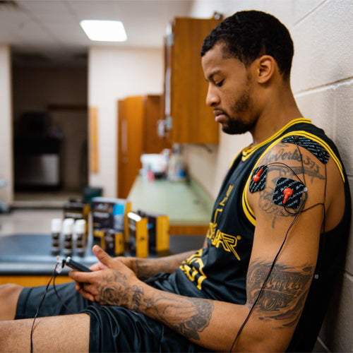 A basketball player in a black jersey sits in a sports therapy room, focused as he uses the iReliev SPORT Muscle Stimulator with Explosive Strength Mode, its electrodes attached to his tattooed upper arm.