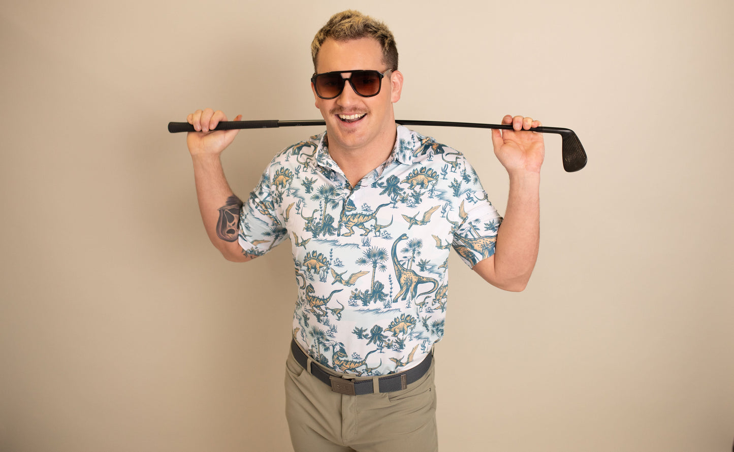 A man with blond hair smiles while wearing Epoch Eyewear's San Juan aviator sunglasses, a dinosaur-patterned short-sleeve shirt, and beige pants. He holds a golf club across his shoulders against a cream background.