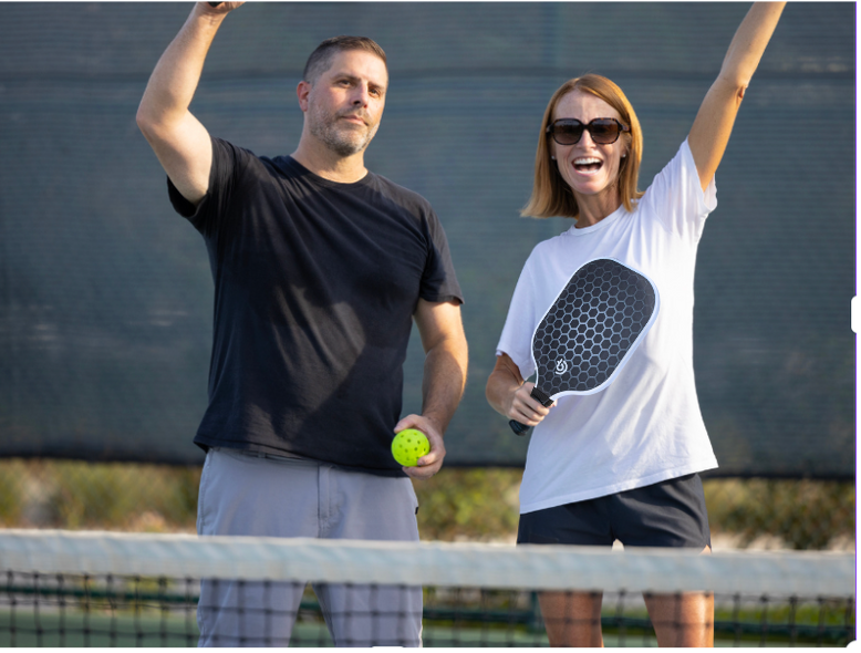 A man and woman holding paddles and a ball stand beside The Big Dink - Portable Pickleball Net by The Big Dink, which is travel-friendly.