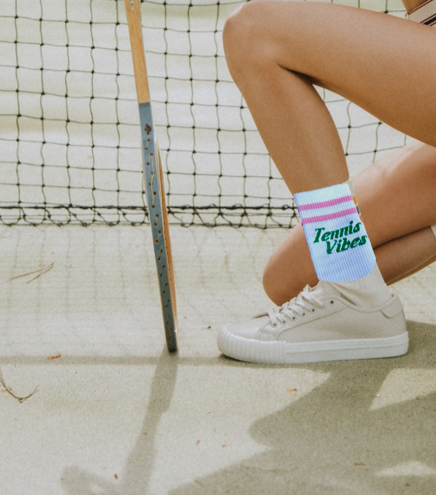On a sunlit court, a person kneels in Runway Athletics' Tennis Vibes Socks—pink, green, and white—with matching white sneakers, a wooden racket, and a black net completing the sporty look.