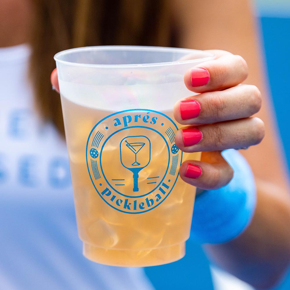 A hand with bright pink nails holds a Swinton Pickleball Aprés Pickleball Reusable Party Cup filled with iced drink. The cup’s blue logo features a cocktail glass and paddles. A blue sweatband completes this perfect pickleball gift!.