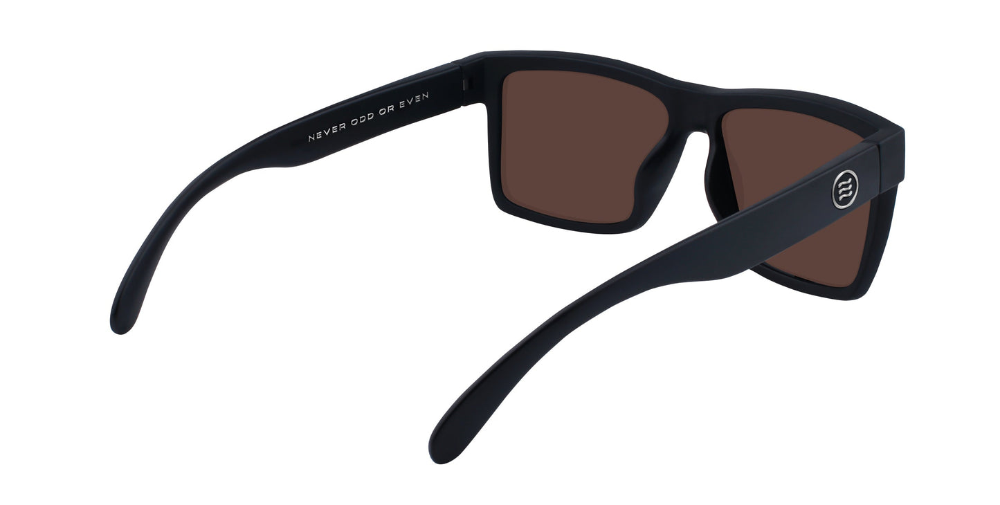 The Montauk by Neven Eyewear features sleek black rectangular frames and brown lenses, shown from a rear angle. The inside left arm displays white text: NEVER ODD OR EVEN—modern eyewear ideal for elevated beach fashion.