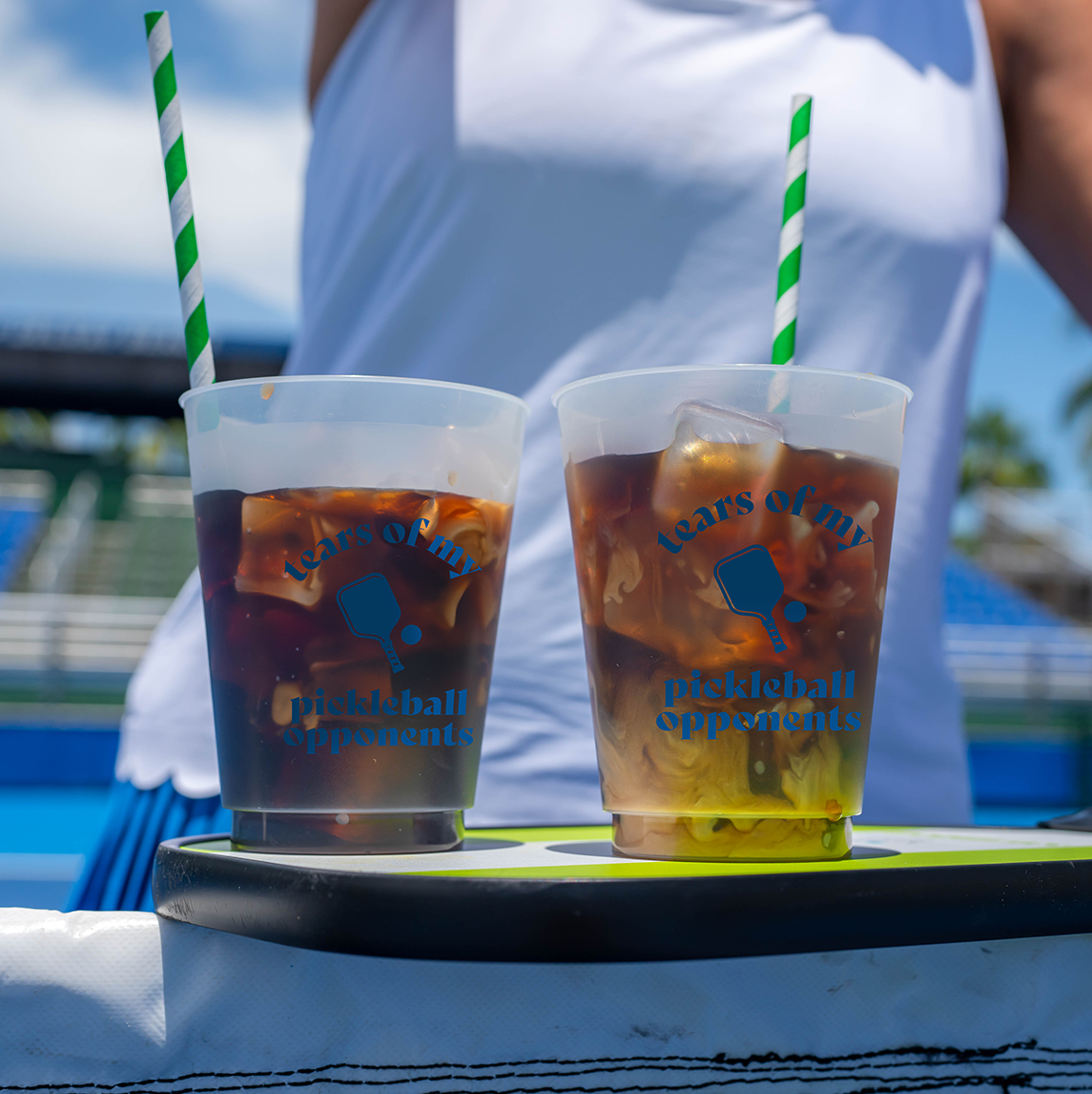 Two Swinton Pickleball "Tears of my Pickleball Opponents" Reusable Party Cups with green striped straws, filled with iced drinks, rest on a paddle—ideal for any sunny court or pickleball party.