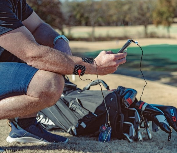 Kneeling on grass beside a golf bag, a person in shorts wears a smartwatch and uses their phone with wired earphones. They use the iReliev SPORT Small Pads Refill Kit for high-performance muscle targeting before play on the golf course.