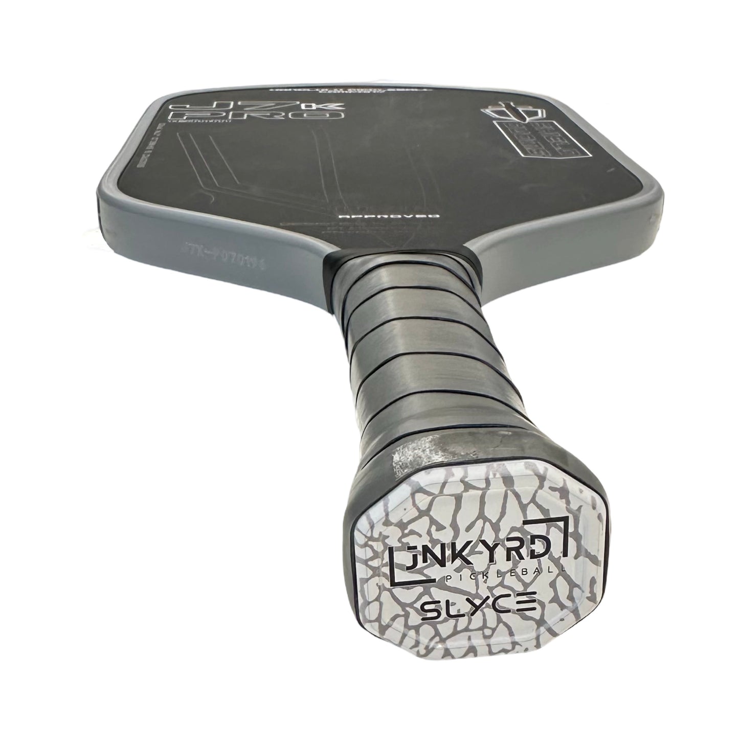 Close-up of the Slyce Sport Special Edition Junkyard Pickleball SpeedCap (28g, 1.0 oz.) Paddle Performance Enhancer, shown from the handle upward with black grip tape and a “JUNKYARD ELITE SLICE” cap over a cracked pattern; paddle face features white graphics.