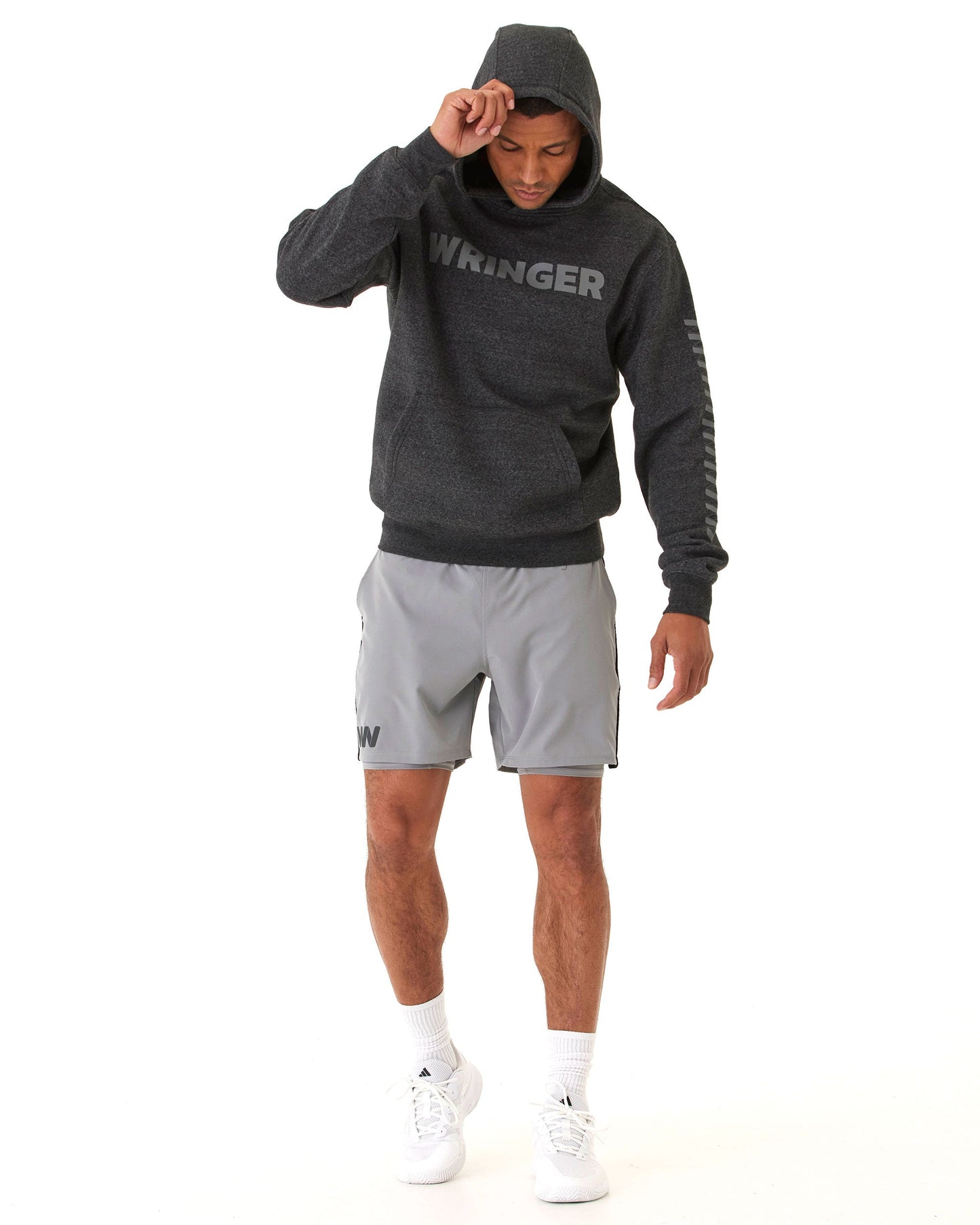 A man stands against a white background wearing a dark gray WRINGER hoodie, Wringer Wear Mojave Performance Shorts 6" Lined in light gray, white socks, and white sneakers. He looks down, adjusting his hood with his right hand.