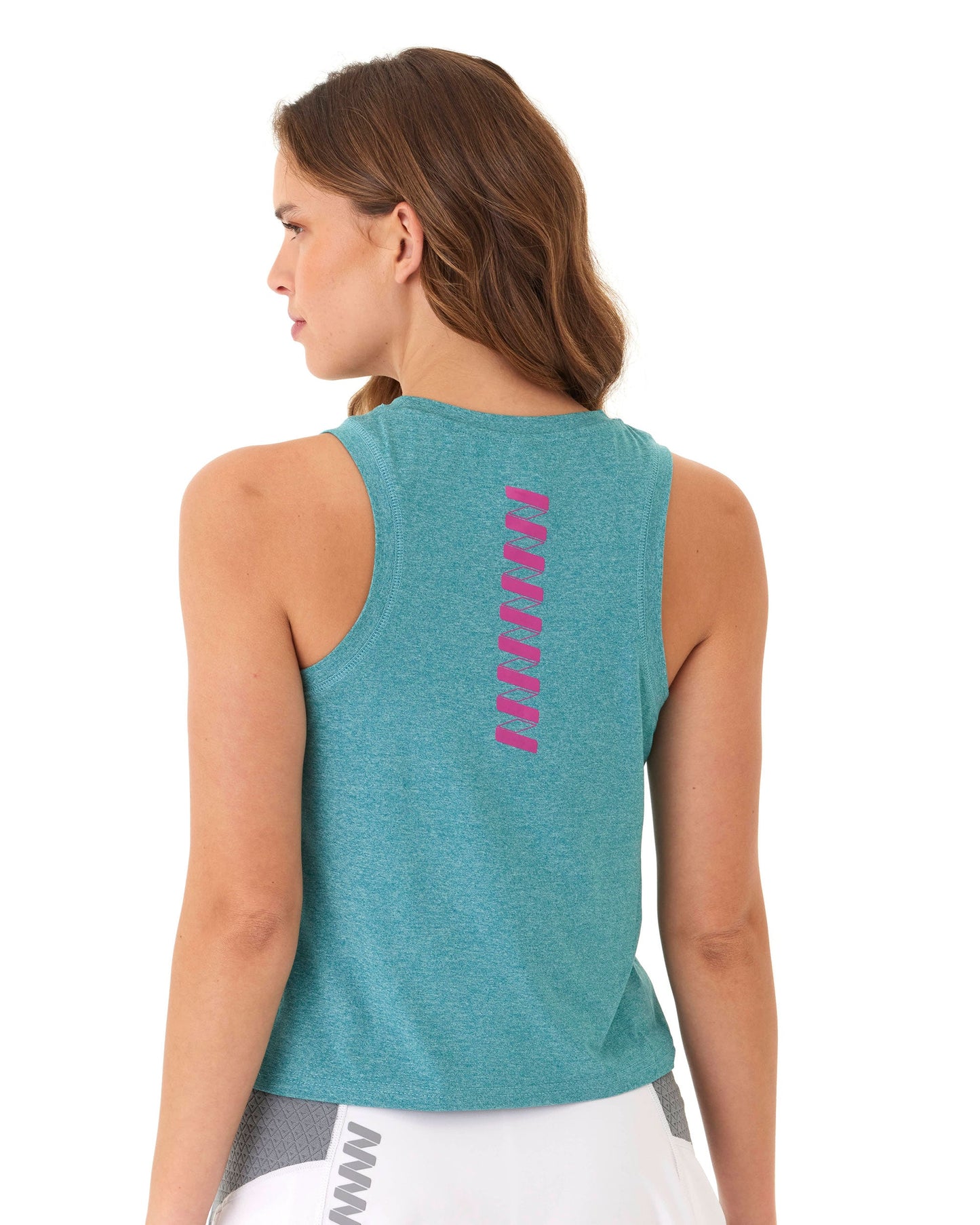 A woman with wavy brown hair models the Wringer Wear Women's Performance Logo Tank in teal with pink zigzag accents on the upper back, paired with white athletic shorts featuring gray mesh panels, against a plain white background. She faces left in profile.