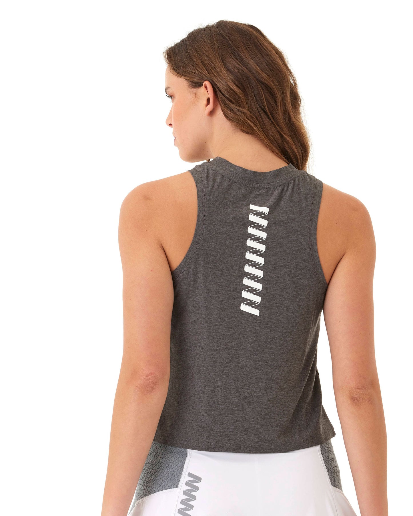 A woman with long brown hair models the Wringer Wear Women's Performance Wringer Tank, a sleeveless dark gray top with a white zigzag detail on the upper back, paired with a white skirt. She faces away against a plain white background.