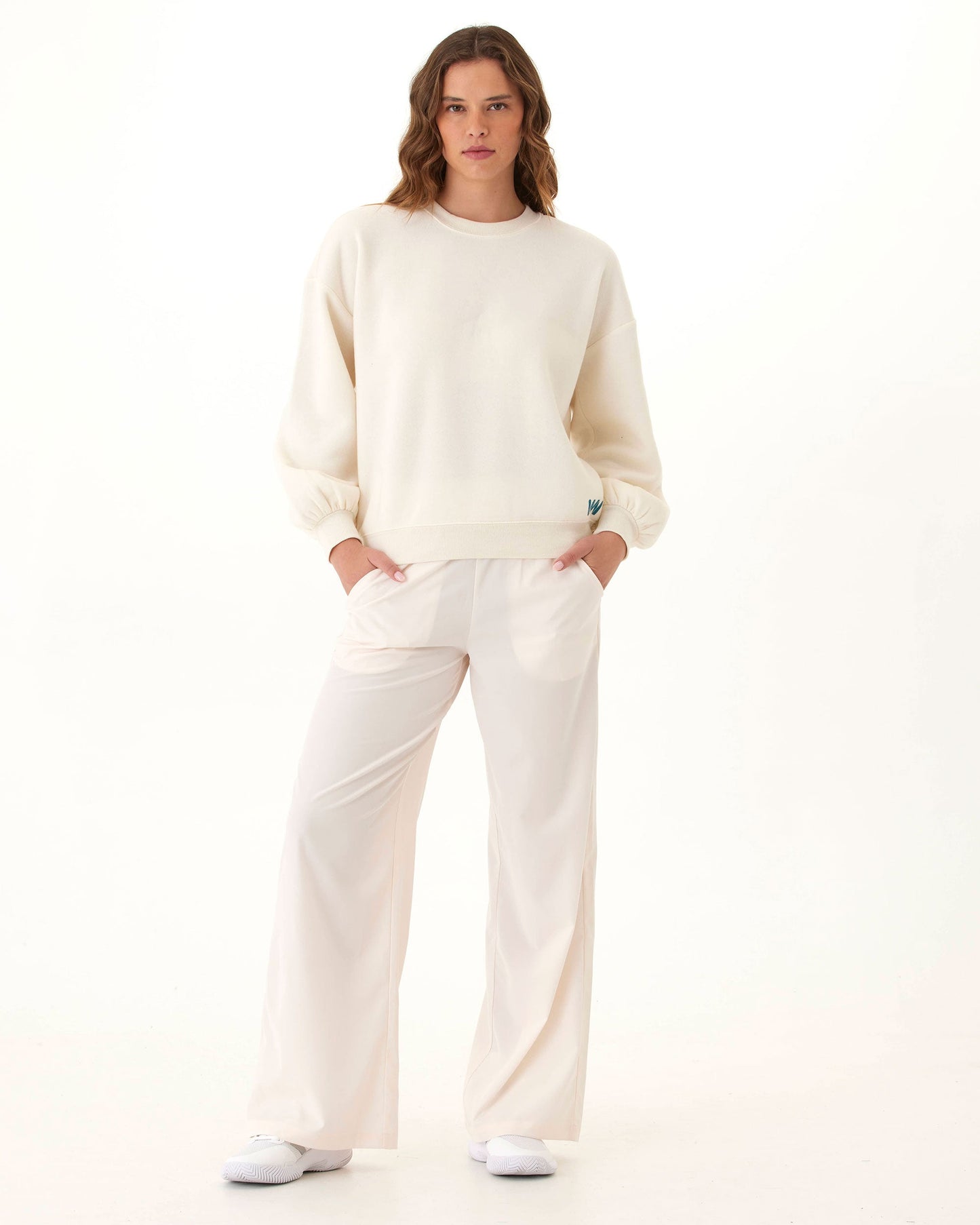 A woman faces forward in Wringer Wear's Women's Wide Leg Pant, made from hydrophobic stretch-woven fabric. With wavy brown hair, white sneakers, and hands in pockets, she showcases a minimal court-to-street style on a white background.
