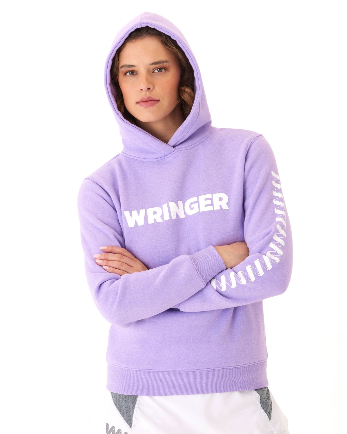 A woman with light skin and wavy brown hair wears the Women's Core Wringer Graphic Hoodie by Wringer Wear in light purple, with bold WRINGER lettering and zigzag sleeves. Its breathable, athletic fit pairs well with her white and gray skirt. Hood up, arms crossed.
