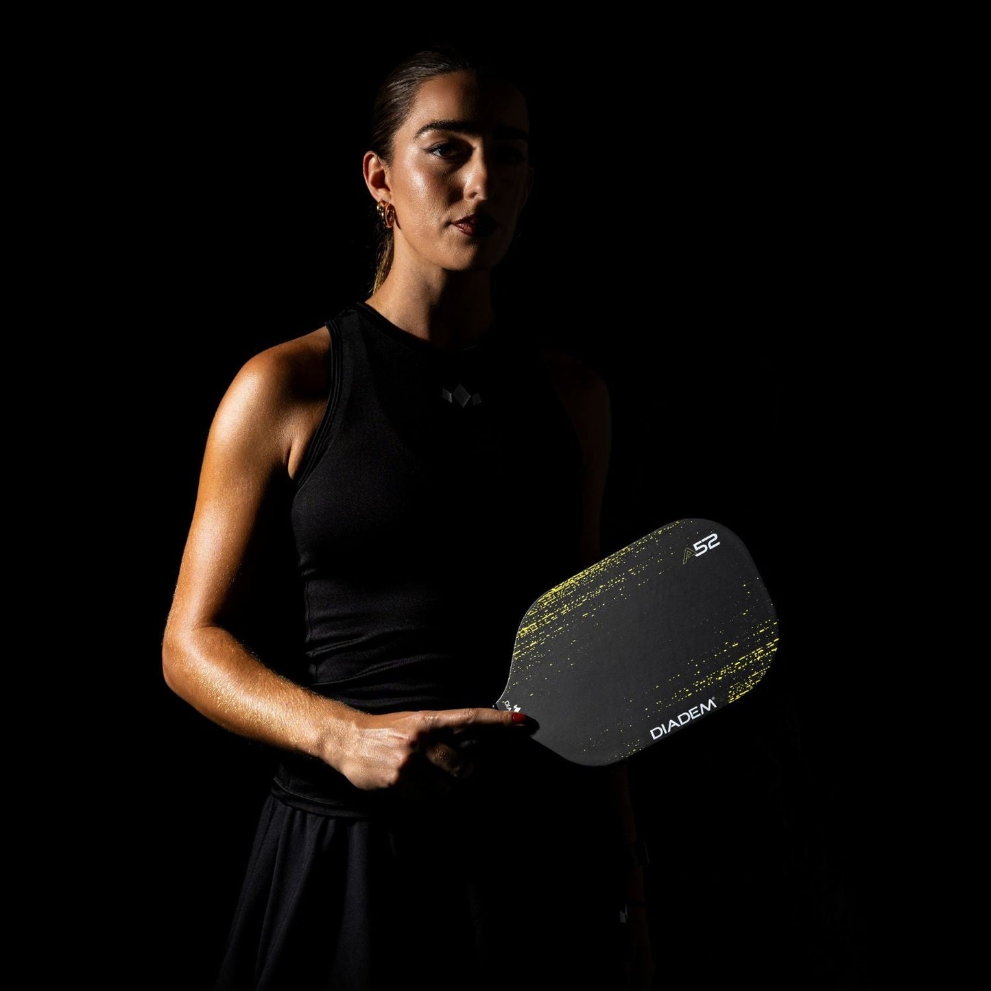 A confident woman stands against a dark background, dramatically lit, holding the Diadem Sports A52 carbon fiber pickleball paddle with yellow streaks. She wears a sleeveless black top and skirt, her hair pulled back as she focuses intently.