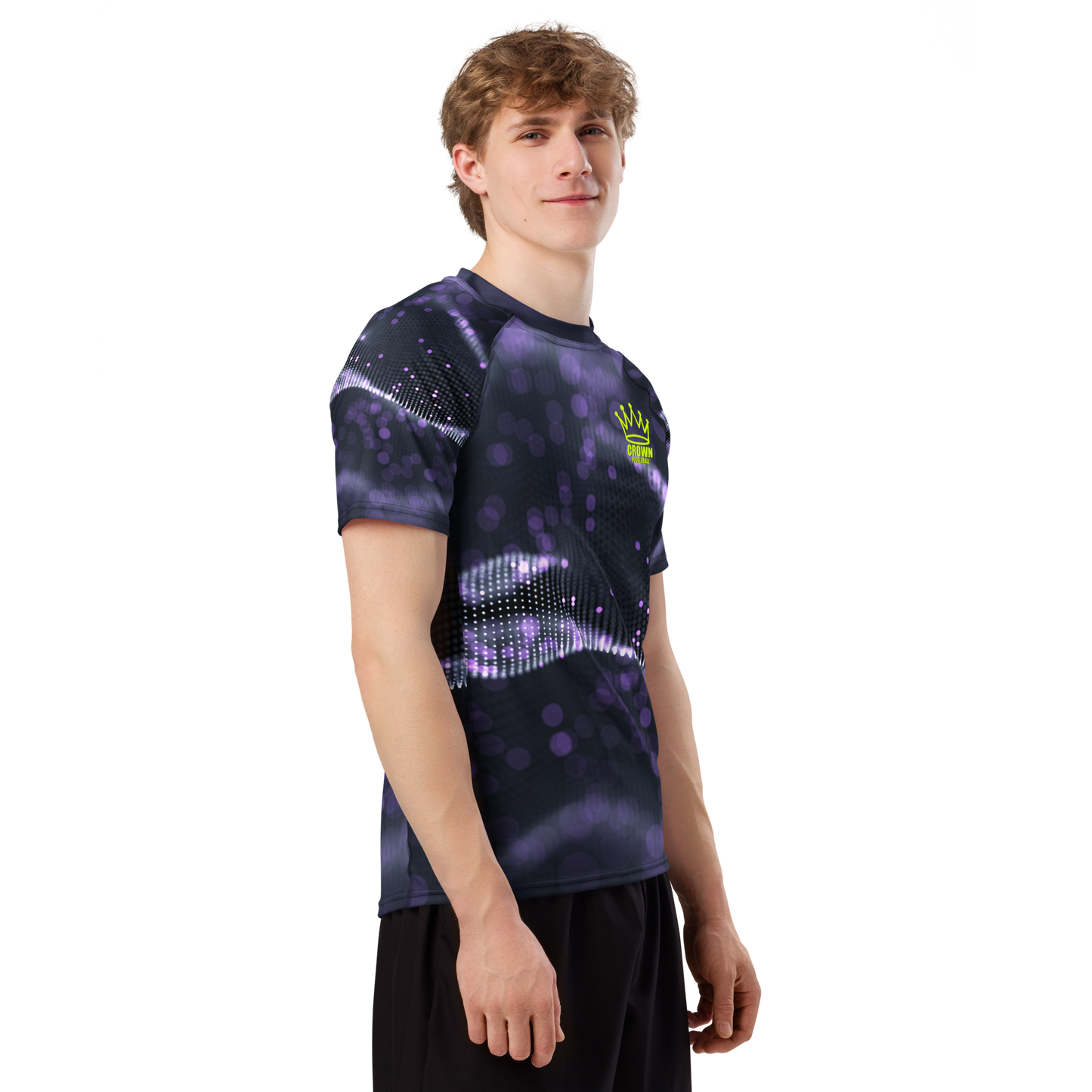 A young man with light brown hair models the Crown Pickleball Men's Tourney Shirt (Purple)—a fitted, moisture-wicking top with a purple and black abstract dotted pattern—paired with black shorts, standing sideways against a plain white background.