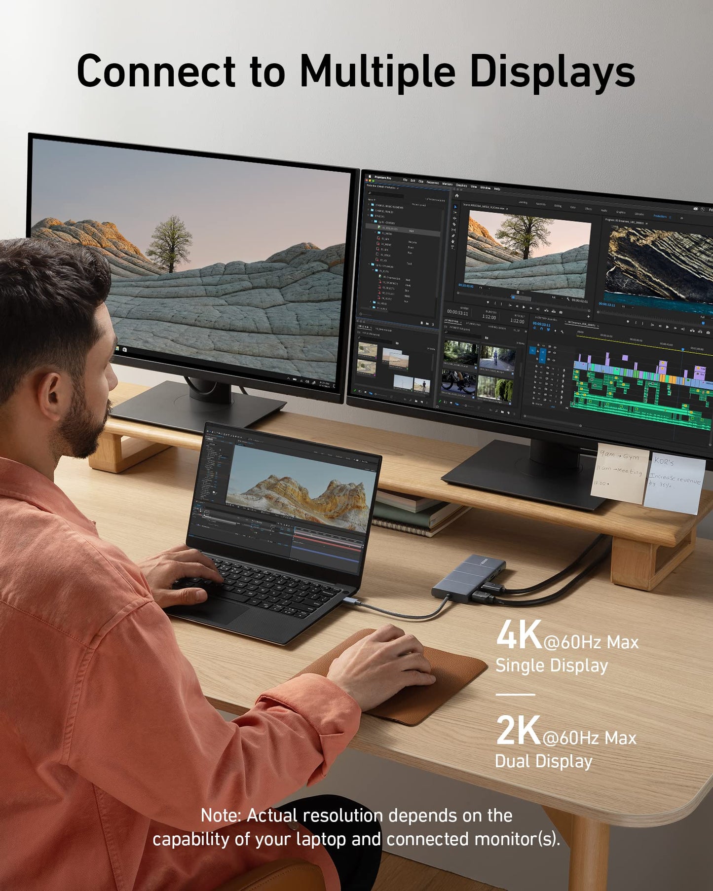 A person uses an Anker 565 USB-C Hub (11-in-1) to connect a laptop to two large monitors displaying creative editing software and landscapes, showing 4K single or 2K dual displays at 60Hz via the 4K HDMI port.