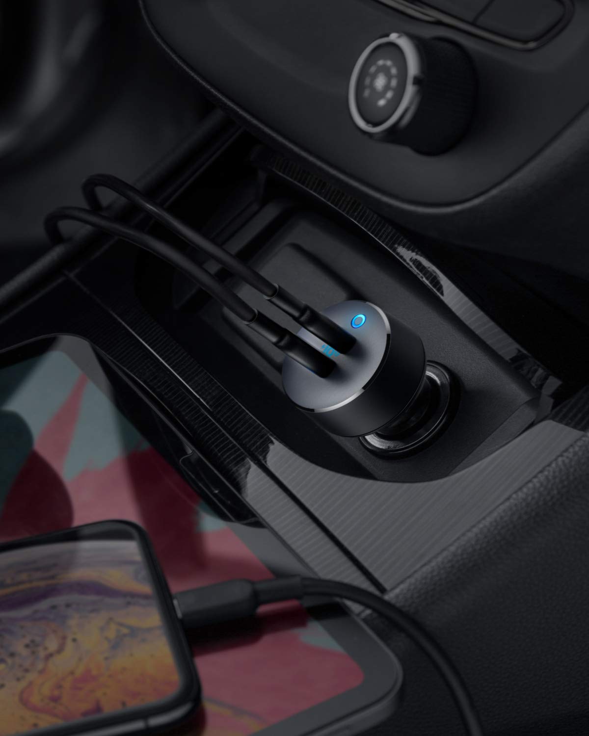 A close-up shows the Anker PowerDrive III Duo USB-C Car Charger with a blue LED light, plugged into a car’s outlet. Two cables connect devices, including a smartphone on the console, for high-speed charging.