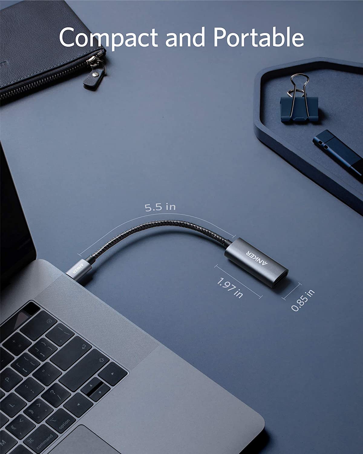 An Anker PowerExpand USB-C to DisplayPort Adapter, measuring 5.5 x 1.97 x 0.85 inches, is plugged into a laptop on a blue desk with a notebook, binder clip, pen, and tray. Text: “Compact and Portable.”.