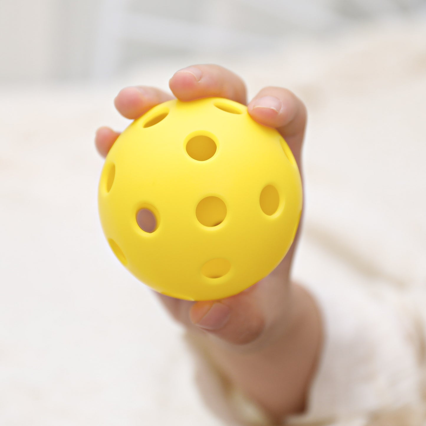 A hand holds the lightweight, bright yellow ARTI Indoor Pickleball by ARTI, featuring evenly spaced round holes. The background is softly blurred and the person’s sleeve is light-colored.