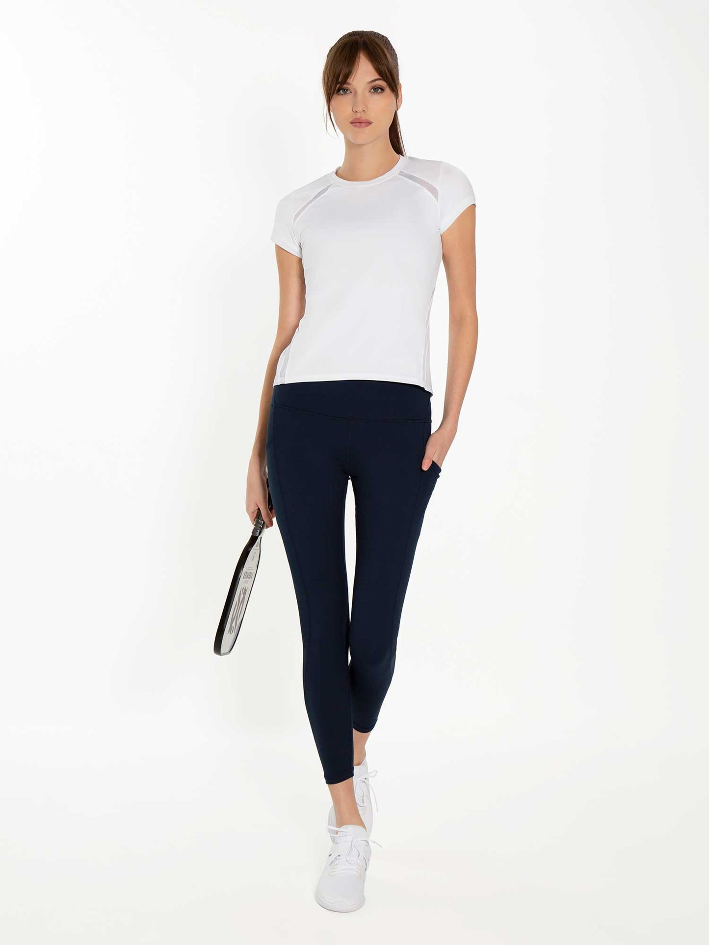 A woman stands facing forward, holding a tennis racket in her right hand. She wears a fitted white short-sleeve top, inPhorm NYC Ashley 7/8 Legging - Midnight with four-way stretch and moisture management, and white sneakers. Her brown hair is in a ponytail.