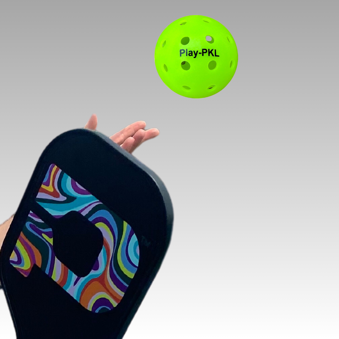 A hand tosses a bright green Banger Hard Surface Pickleball by Playly Store—perfect for indoor courts. In the foreground, a paddle with a colorful wavy pattern is ready to strike against a smooth gray background. Available in a 3-pack.