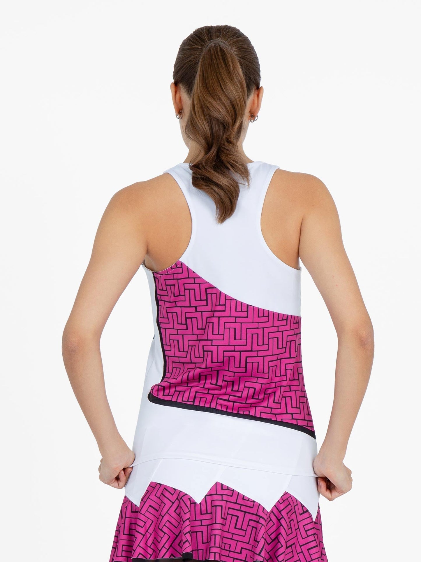 A woman with a low ponytail faces away, wearing the inPhorm NYC Baseline Tank in White/Pink Tessel and a matching skirt, holding the hem of her moisture-wicking top with both hands.