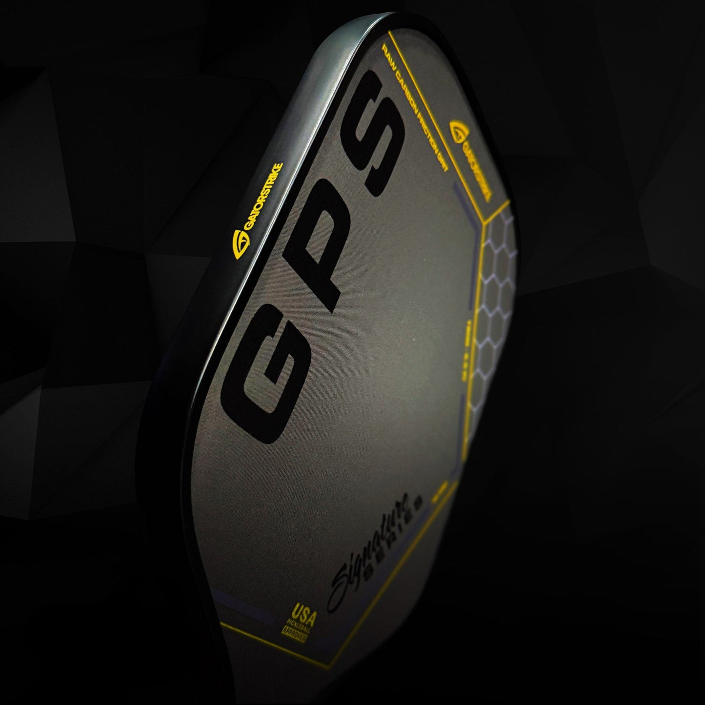 A close-up of the GATORSTRIKE™ Gold Pro Series black carbon fiber pickleball paddle by GATORSTRIKE, featuring bold GPS letters, yellow accents, hexagonal patterns, and anti-vibration foam against a dark abstract background.