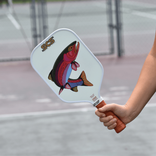 A person holds a Salted City Sports Evo-Lite Series Bow pickleball paddle with a brown grip and colorful fish graphic, marked "SCS" near the top. The USA Pickleball approved paddle appears against a blurred outdoor sports court.