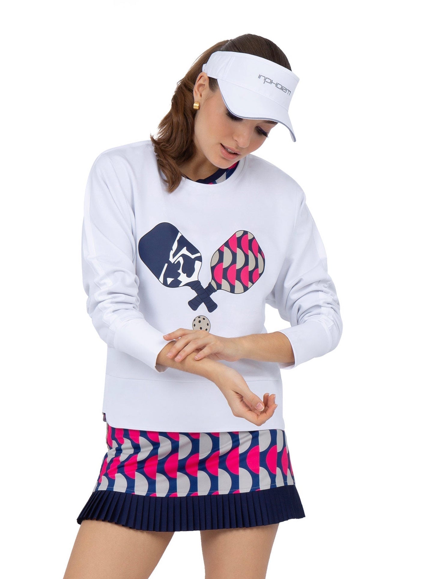 A woman in the Camila Performance Sweatshirt - Pickleball Print by inPhorm NYC, made from UPF 50+ performance fabric, pairs it with a colorful pleated skirt and white visor, showcasing stylish court-ready comfort.