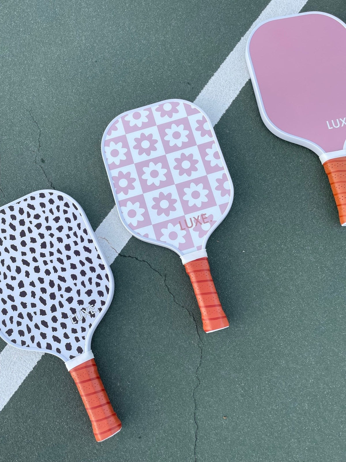 Three LUXE Pickleball "Checkered Flower" USA Pickleball Approved paddles with orange grips sit on a green court near a white line. Designs shown include black/white leopard, pink floral, and solid pink.