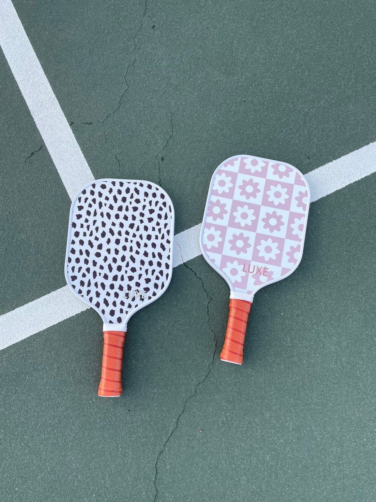 Two LUXE Pickleball paddles with orange grips lie on a green court. The left is the Cheetah model in cheetah print; the right features a pink floral checkerboard. White lines cross beneath them.