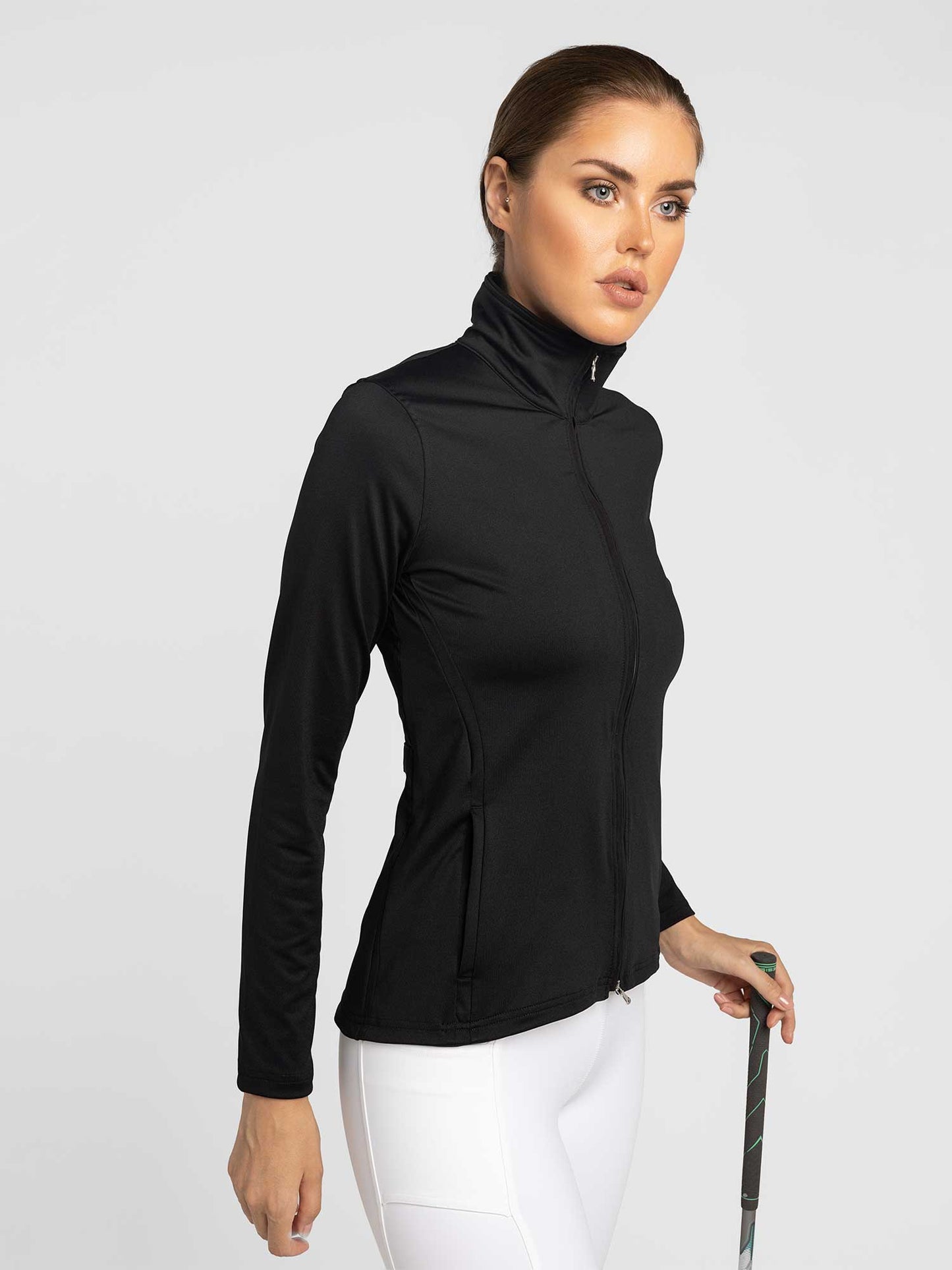 A woman with brown hair tied back models the inPhorm NYC Classic Blythe Jacket in Black and white leggings, holding a golf club with her left hand and gazing ahead with determination against a plain light background.