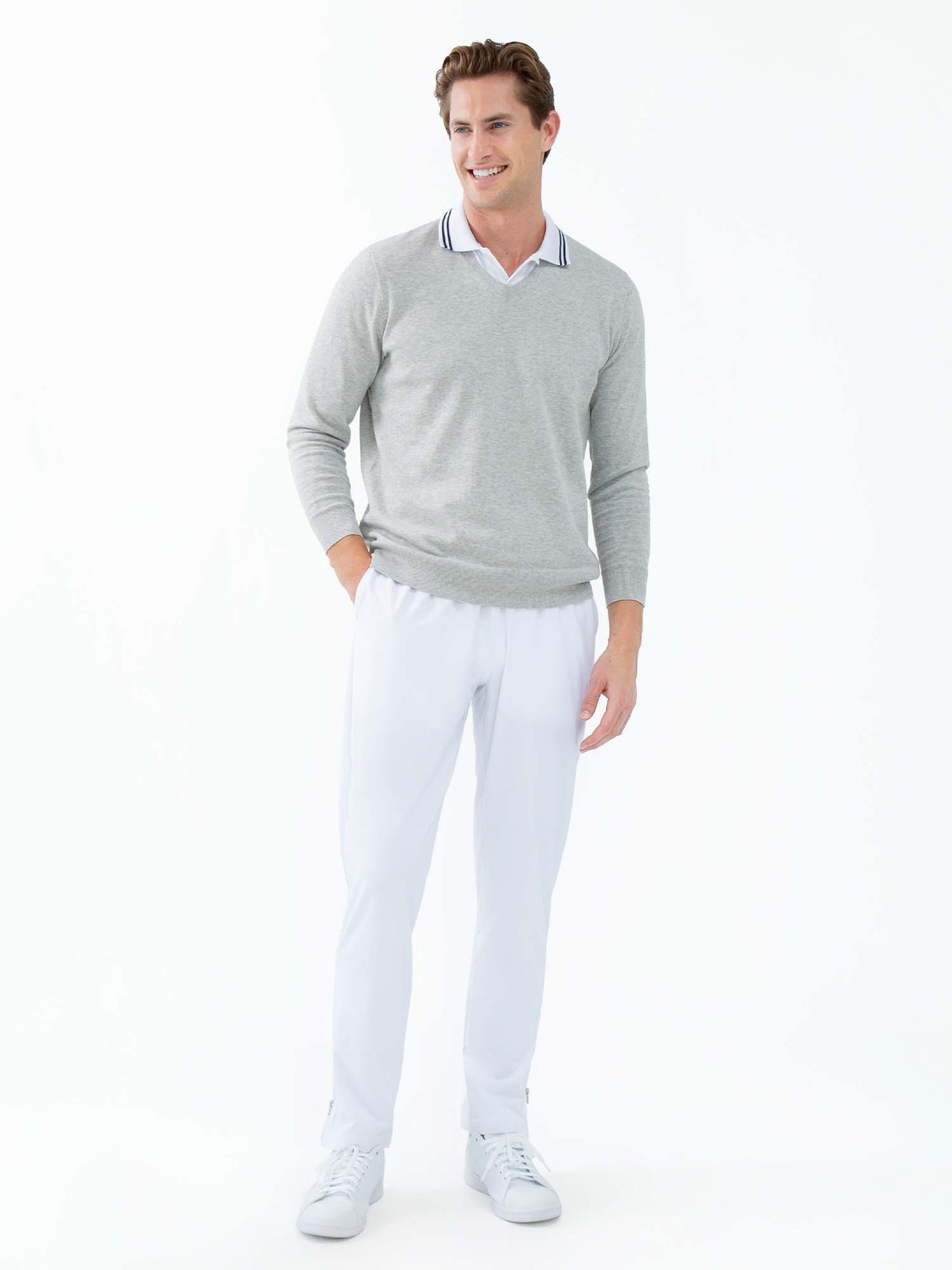 A man smiles against a white background, wearing the inPhorm NYC Classic Performance Men's Tennis Pant in white, paired with a light gray sweater over a white collared shirt. His right hand is in his pocket; his left arm rests by his side.