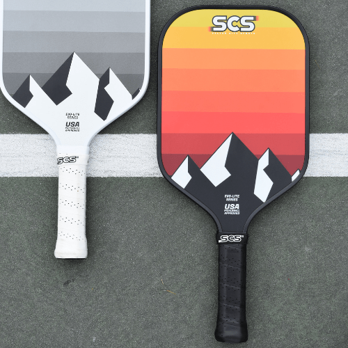 Two Salted City Sports Evo-Lite Series paddles lie on the court: left is a grayscale mountain pro paddle, right has a Utah Jazz-inspired red, orange, and yellow gradient. Both are USA Pickleball approved with black handles and SGS branding.