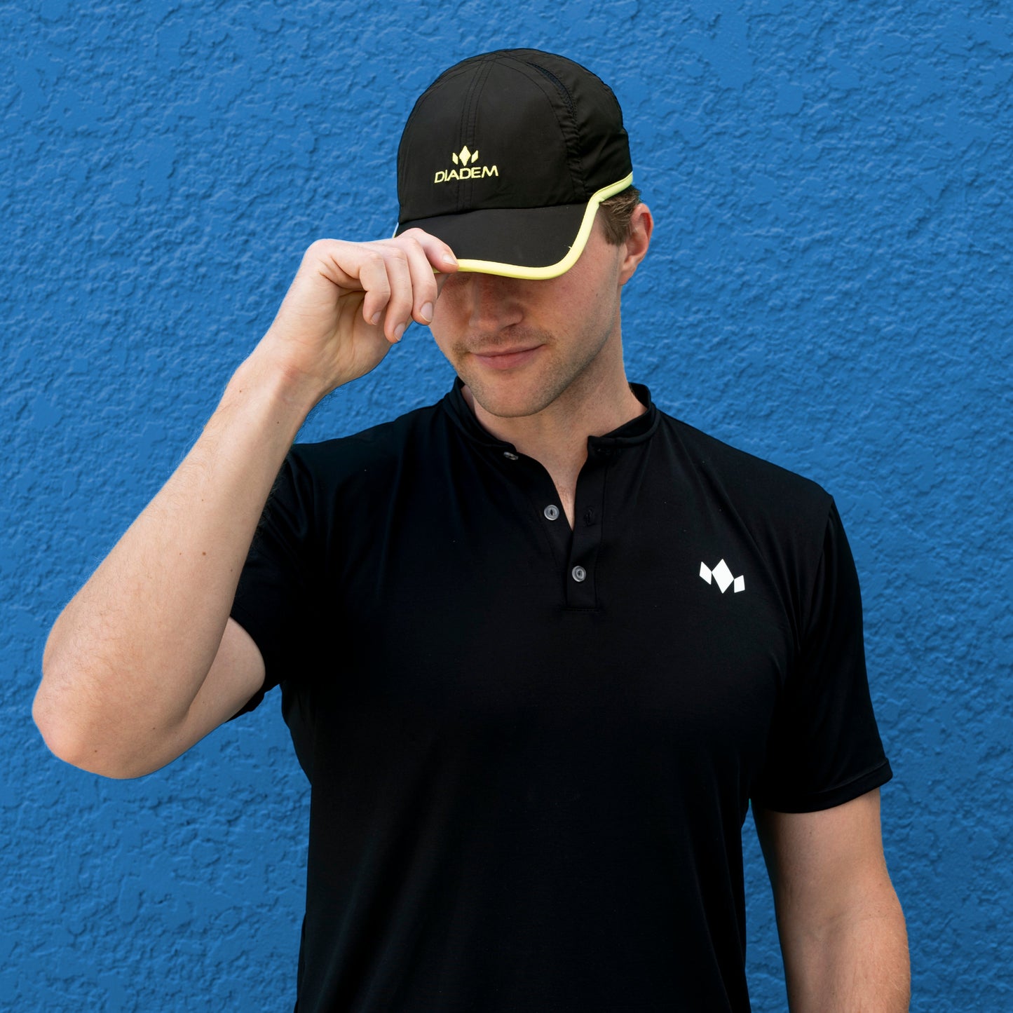 A person in front of a blue textured wall wears a black polo and a Diadem Sports Performance Hats Snapback, both featuring the white and yellow Diadem Sports logo. The yellow-trimmed cap is tilted to partially cover their face.