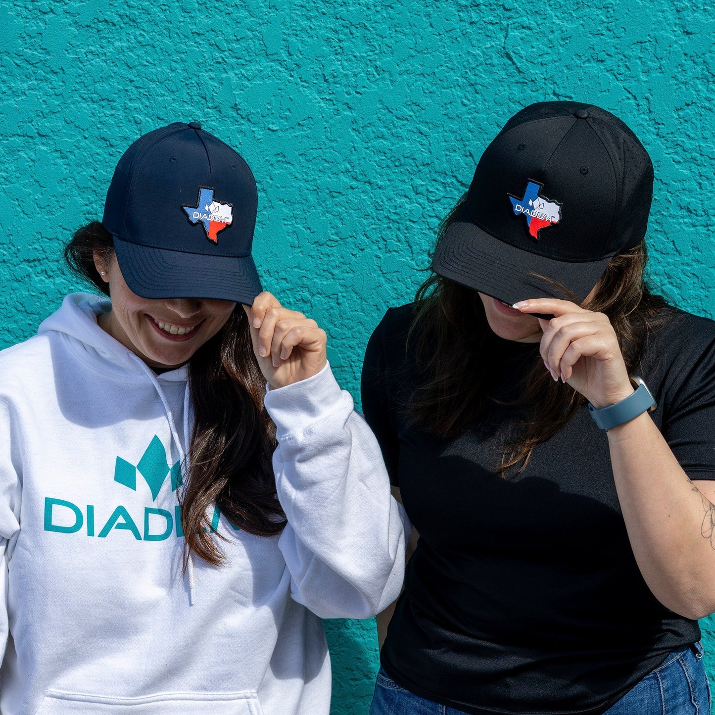 Two people pose against a teal wall, both holding the brims of their Diadem Texas Performance Hats by Diadem Sports. The left wears a white "DIADIA" hoodie, the right a black shirt and blue smartwatch. Their faces are partly hidden.