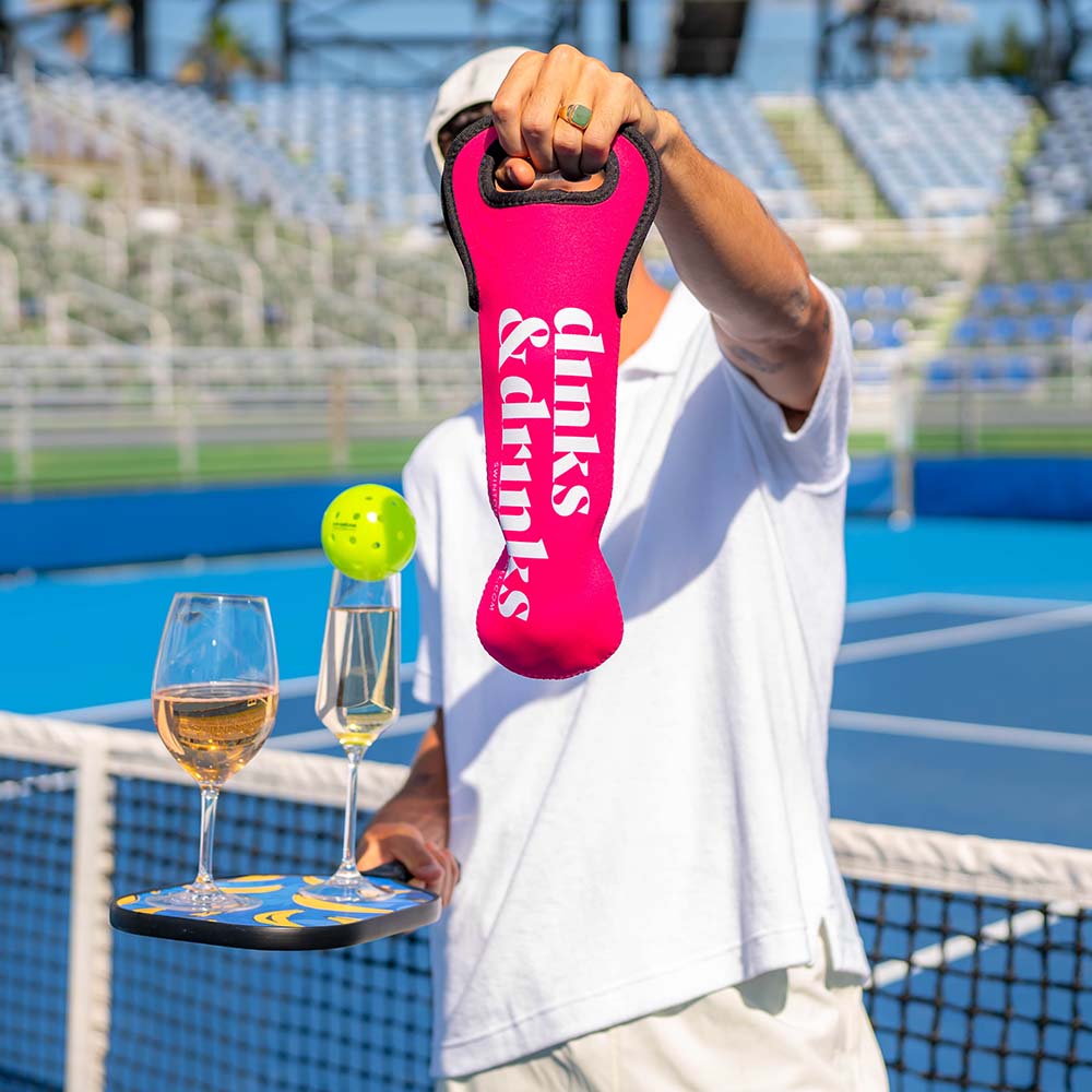 A person in a white shirt holds Swinton Pickleball’s Dinks 'N Drinks Sleeve of Pickleballs on a blue court, while balancing two glasses of white wine and a paddle with a yellow pickleball in their other hand.