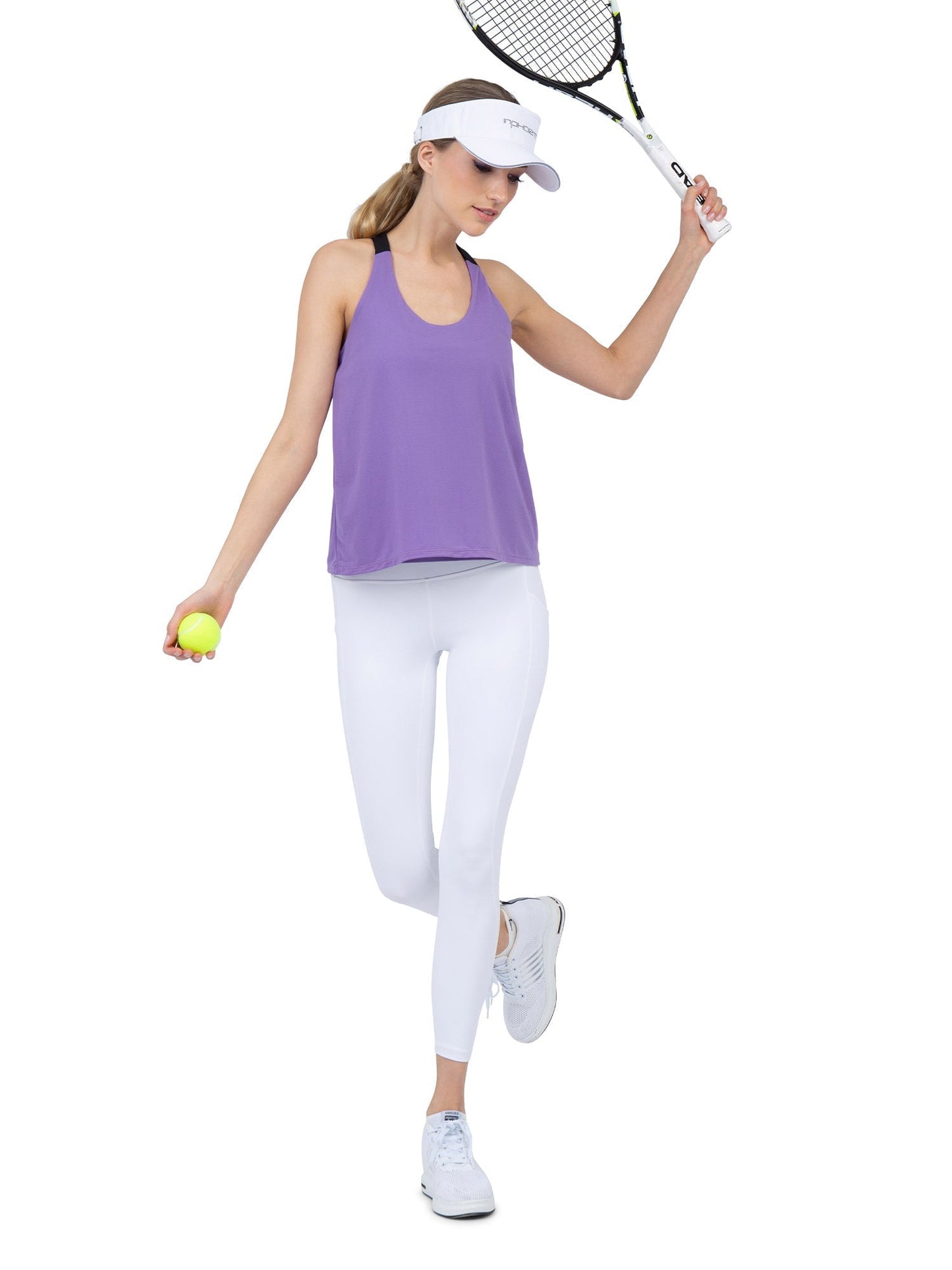 A woman in the inPhorm NYC Double-Layered "T" Racer Back Camila Tank in Lavender/Black stands on one foot against a plain white background, holding a yellow tennis ball and raising a racket while looking down.