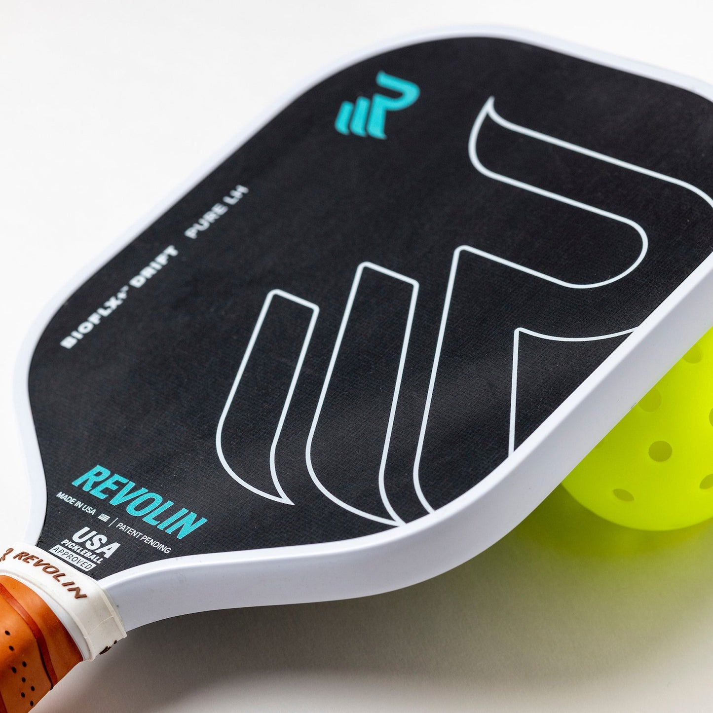 A close-up of the Revolin Sports Drift Pure LH Pickleball Paddle in black with white and turquoise graphics, honeycomb core for reduced vibration, orange-brown grip, and white edge, overlapping a bright yellow pickleball on a white background.