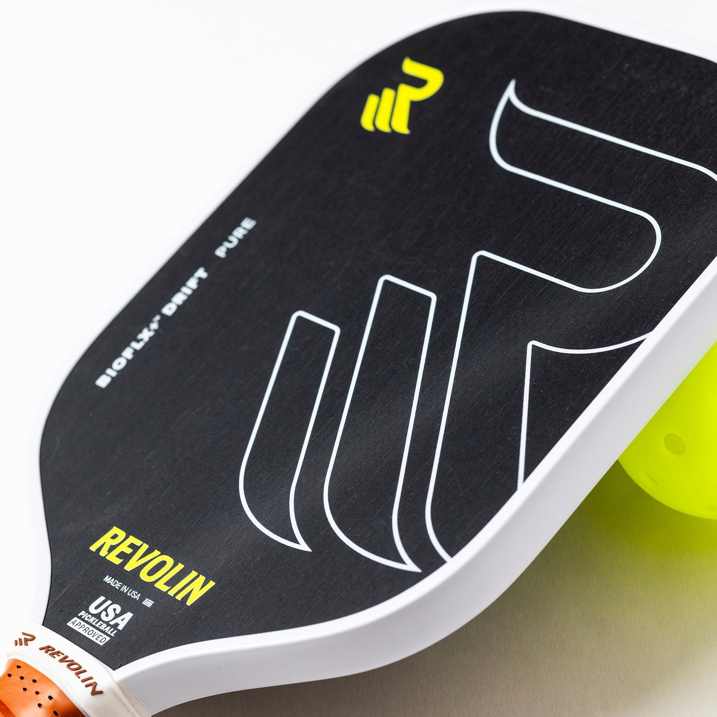 Close-up of a black Revolin Sports Drift Pure Pickleball Paddle with white and yellow accents, bold geometric design, and “REVOLIN” in yellow. An orange handle and a yellow pickleball are beside the paddle for enhanced control.