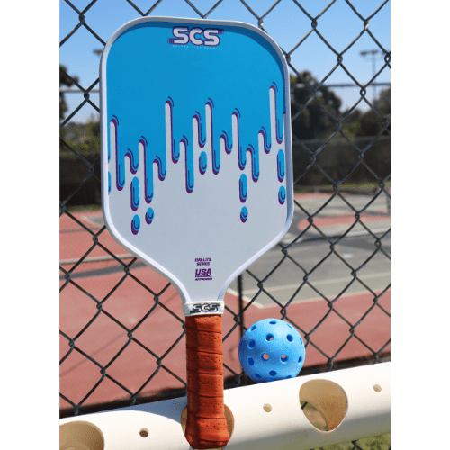 A blue and white Salted City Sports Drippy | Evo-Lite Series pickleball paddle with a brown grip leans on a chain-link fence near tennis courts. USA Pickleball approved, it offers excellent spin control. A blue pickleball rests nearby.