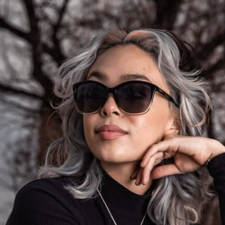 A young woman with wavy silver hair and dark roots wears Epoch Eyewear's Elizabeth luxury sunglasses and a black top, resting her chin on her hand with a slight smile as out-of-focus tree branches fill the background.