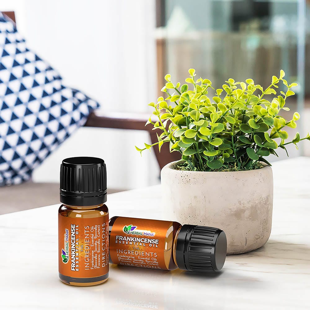 Two amber bottles of Herbal Mana SALE!! Frankincense Essential Oil (Boswellia Blend) sit on a white surface next to a small green potted plant, with a blue and white pillow in the blurred background suggesting a cozy, relaxing space.