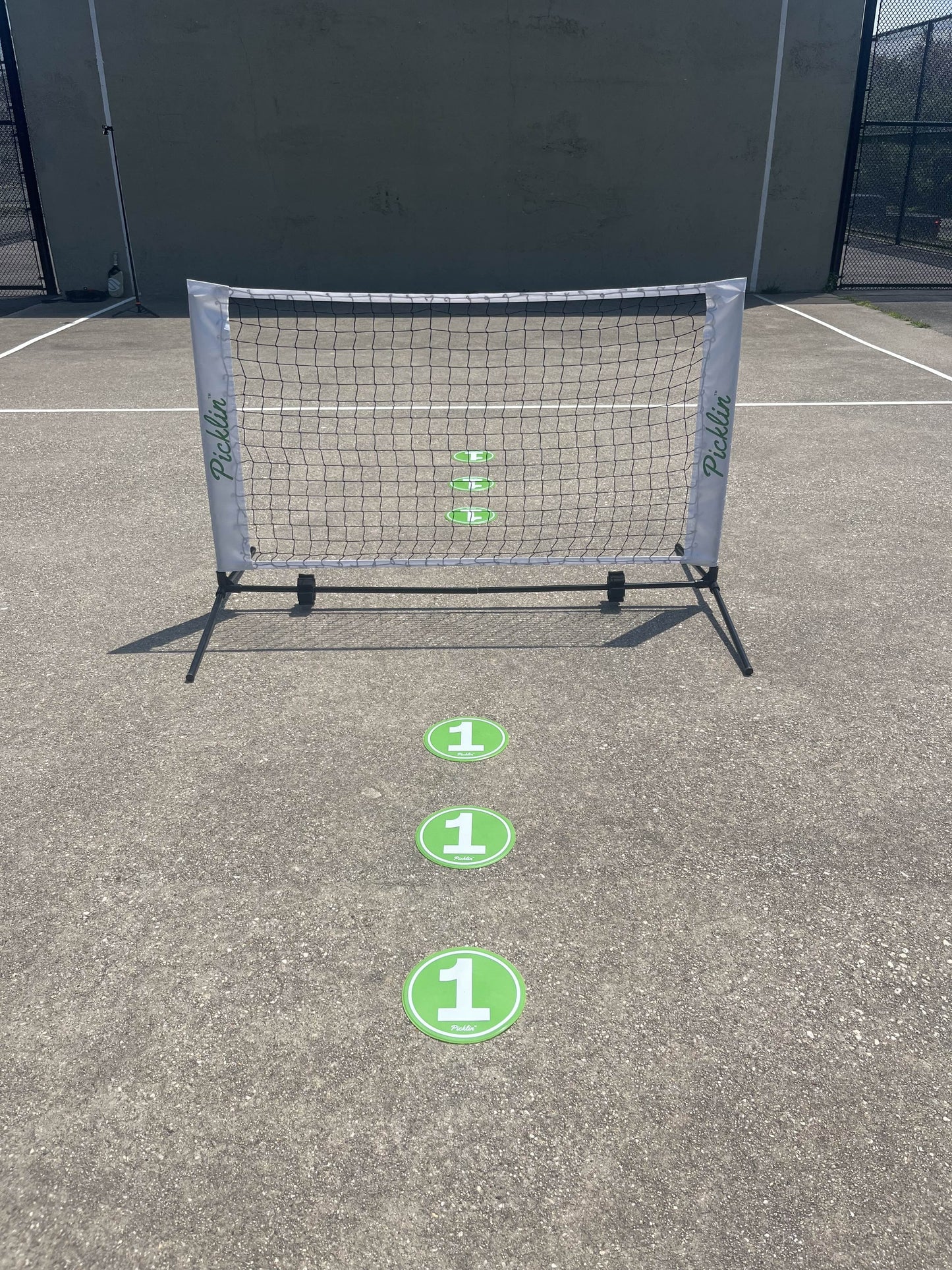 A small Picklin™ Floor Targets set is arranged on a gray outdoor court before a net, with four green targets marked "1" in a line—ideal for training. The scene features a concrete wall backdrop.