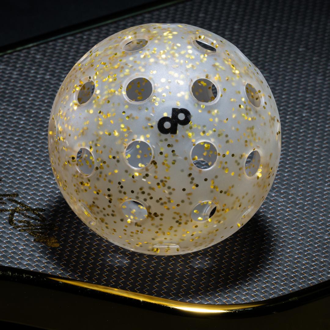 A Gold Glitter-Infused Pickleball Ball by Dope Pickleball Co. from the High Roller 4-Pack sparkles on a dark surface, featuring multiple holes, gold glitter interior (GlitterTech), and a black infinity symbol highlighted by overhead light.