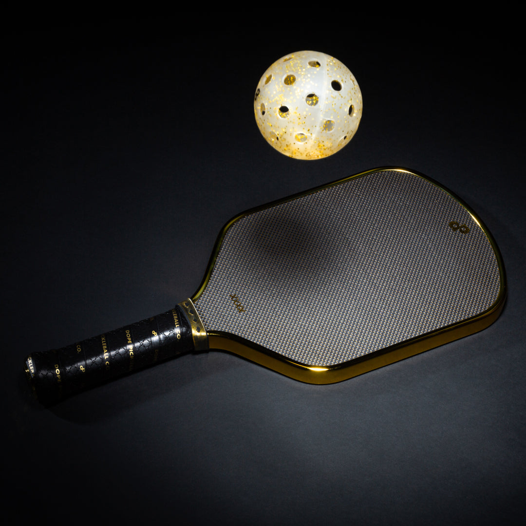 The Dope Pickleball Co. High Roller Pickleball Paddle – The Original Chrome Gold Pickleball Paddle – lies on a dark surface, while a glowing pickleball above it casts brilliant patterns and highlights its chrome gold edges.