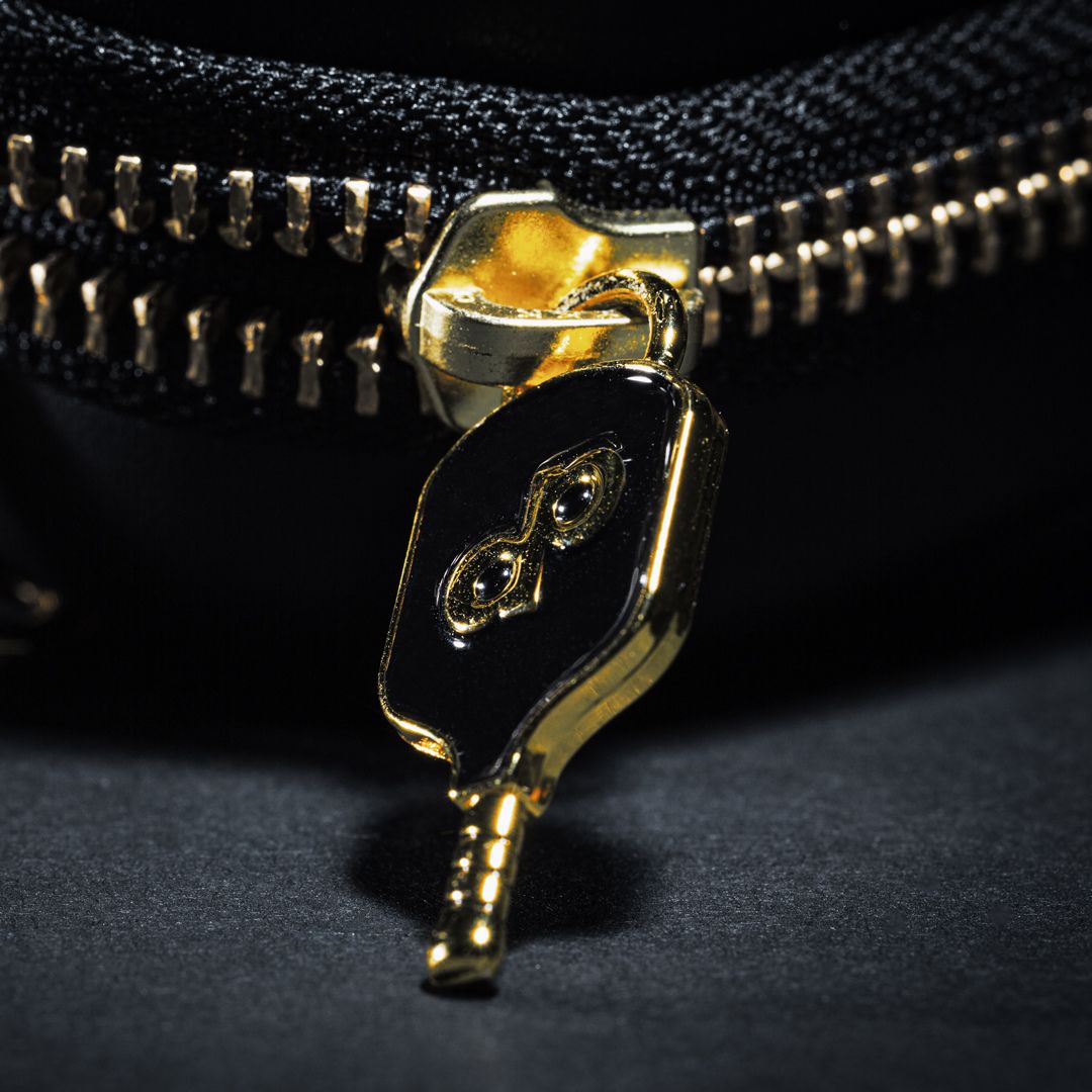 A close-up shows the gold paddle-shaped zipper pull with an embossed infinity symbol on the Dope Pickleball Co. High Roller Pickleball Paddle Travel Case, made of black vegan leather. The dark background highlights its shiny metallic details.