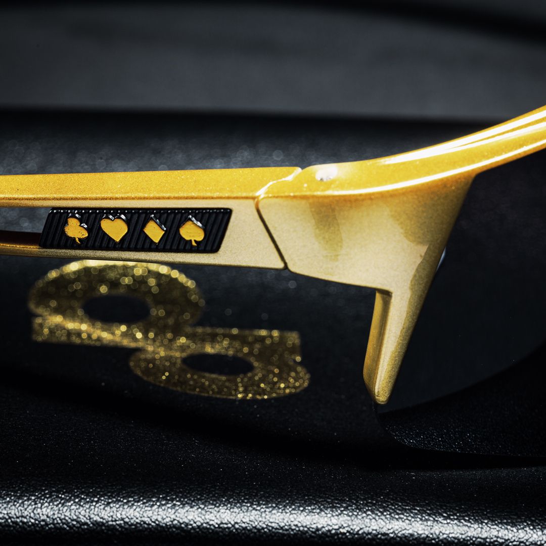 Close-up of Dope Pickleball Co.'s High Roller Performance Pickleball Sunglasses, showcasing gold frames, black arms with card suit symbols, and lenses reflecting glittery gold 88s on a dark background.