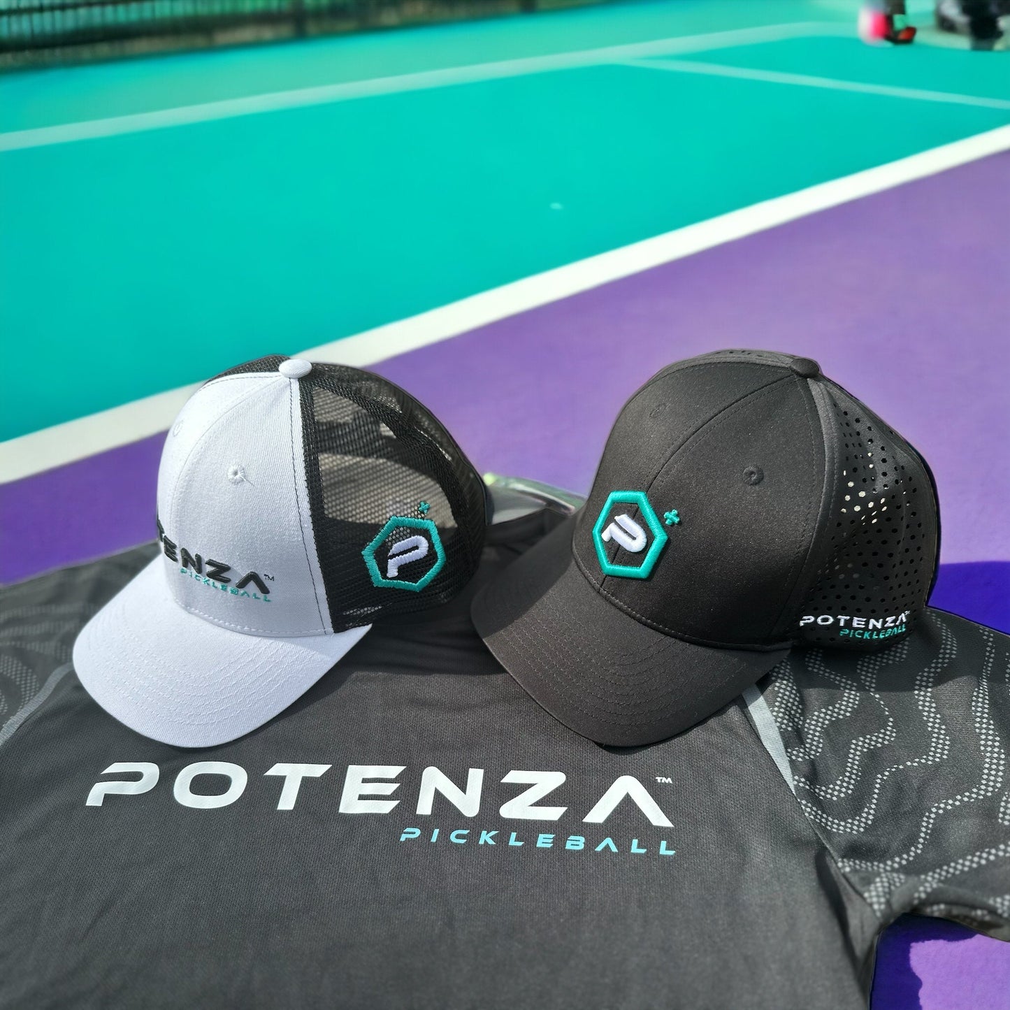Two Potenza Pickleball Performance PRO Hats—one lightweight black, one white/gray—sit on a matching shirt. All items display the Potenza logo and text on a purple and teal pickleball court under bright sunlight.