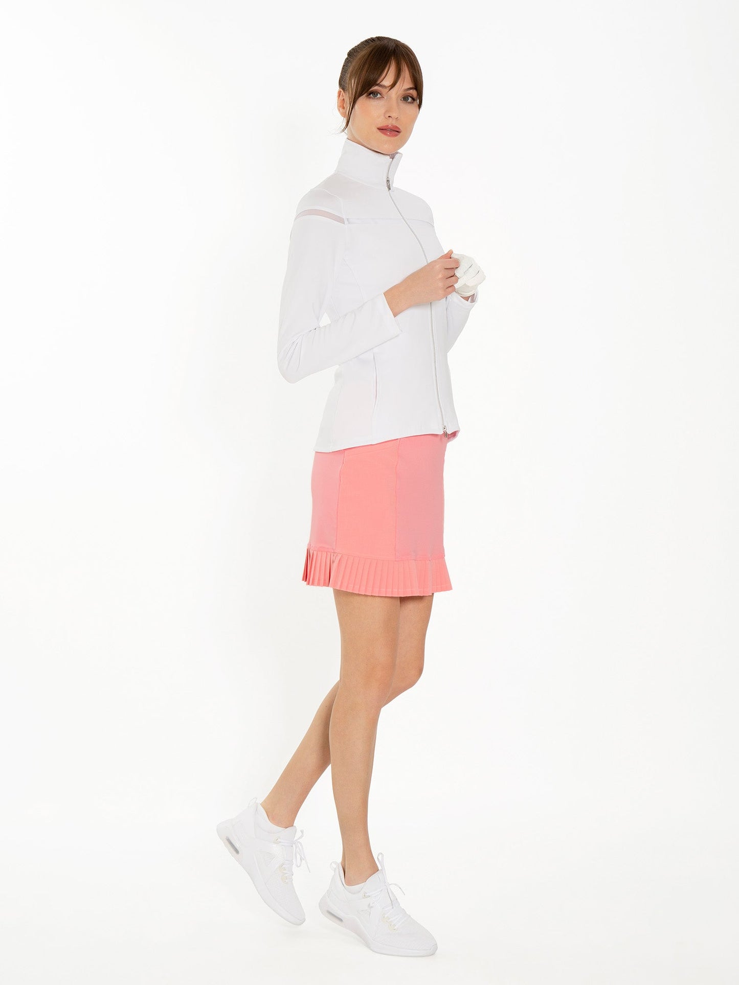 A woman with light skin and brown hair in a low ponytail wears the inPhorm NYC Kai 17" Pleated Skirt in Plumeria, a fitted white zip-up jacket, and white sneakers. She holds a white glove in her left hand against a white background.